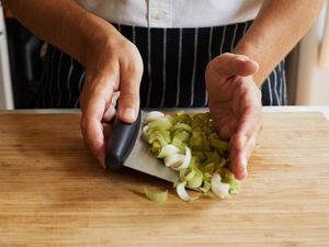A person using a metal bench scraper to scoop up sliced leeks.