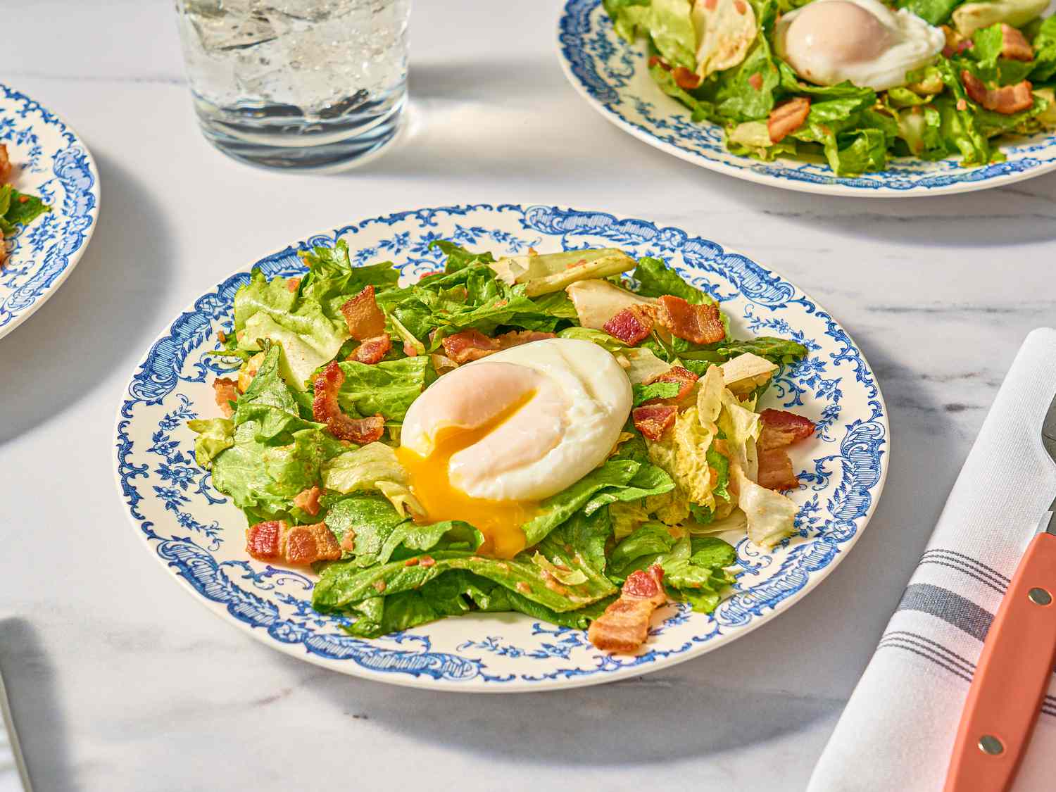 Salade Lyonnaise with broken egg on antique blue and white plates with sparkling water, and utensils on white and grey striped napkin on a white marbled surface