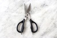 A pair of Cangshan 9-inch Heavy-Duty Come-Apart Kitchen Shears with black handles and serrated blades placed on a marble surface