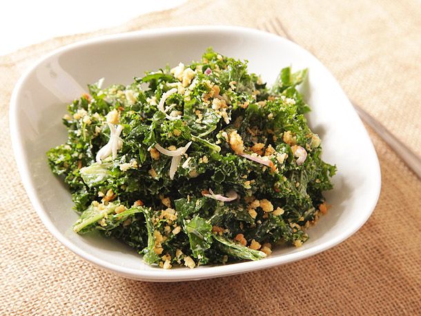 A close-up of kale caesar in a small white bowl, set atop a beige place setting.