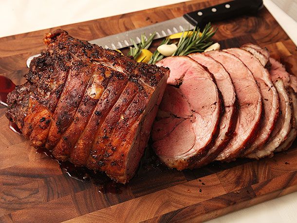 Finished lamb roast sliced and resting on a cutting board, with a carving knife and rosemary and garlic next to it.