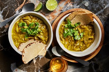 2 bowls of Chicken Chile Verde with ripped tortillas, limes and cilantro, on black marble surface