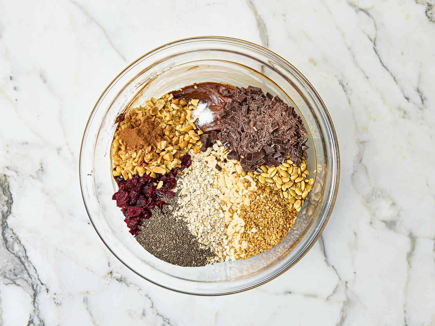 A bowl containing various ingredients for homemade protein bars including oats seeds dried cranberries and chocolate shavings