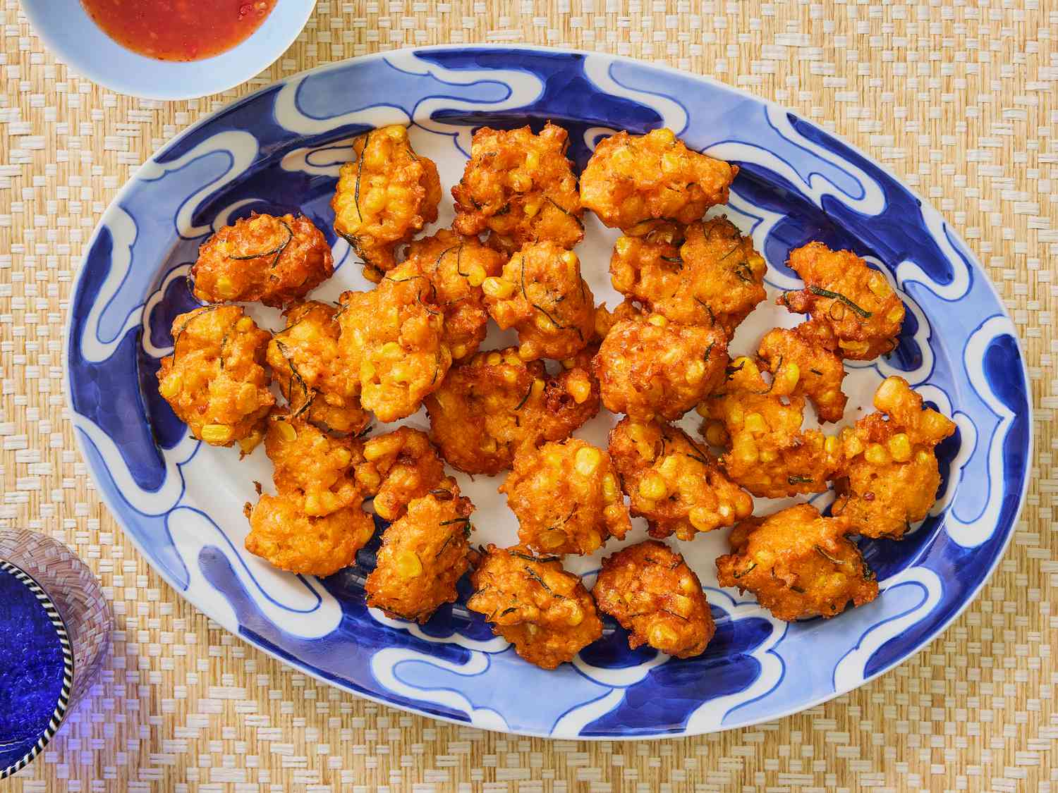 Fritters on a blue and white printed plate, on a bamboo mat. Glass of blue water, and red chili sauce on the side 