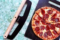 A pepperoni pizza on a cutting board next to a Gozney pizza rocker with a wooden handle