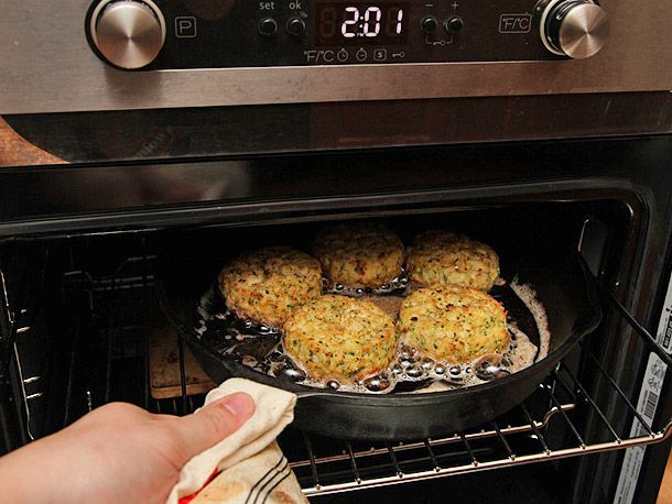 Placing a cast iron skillet containing five crab cakes into the oven to broil. 