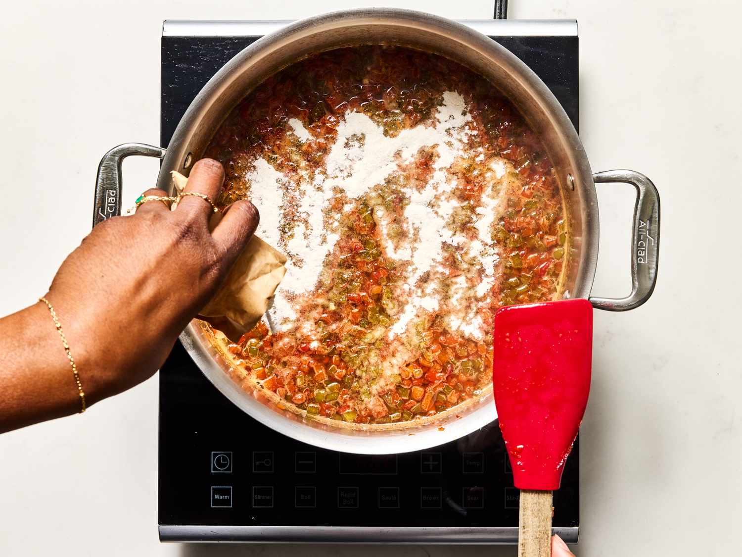 A hand pouring liquid into a pot of simmering ingredients on a stovetop, with a red spatula resting on the side of the pot