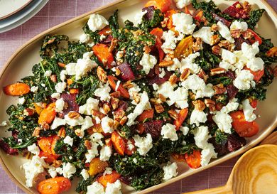 A salad made with kale roasted root vegetables and crumbled cheese topped with nuts on a rectangular serving dish