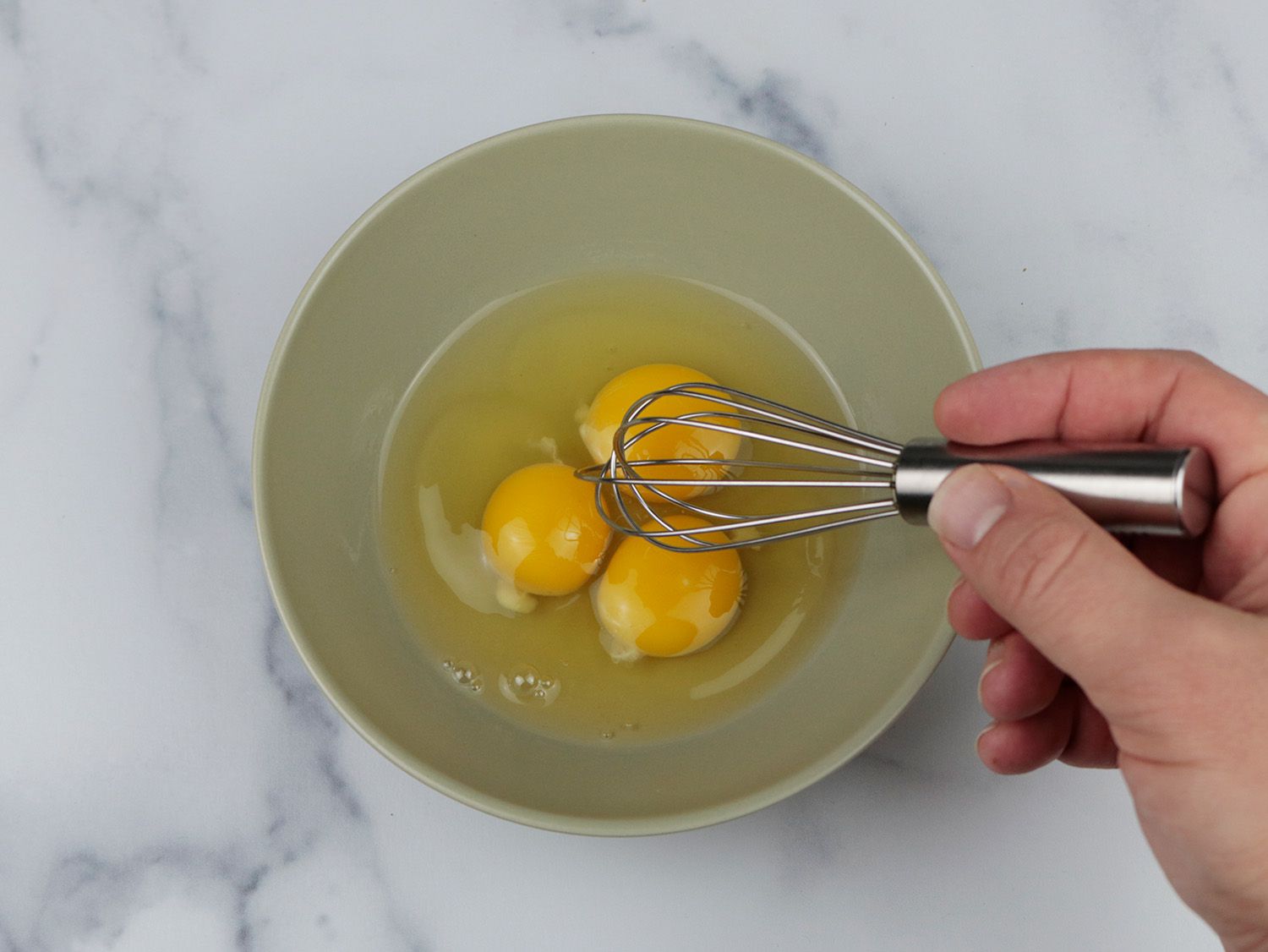 a miniature whisk and three eggs in a small bowl on a marble surface