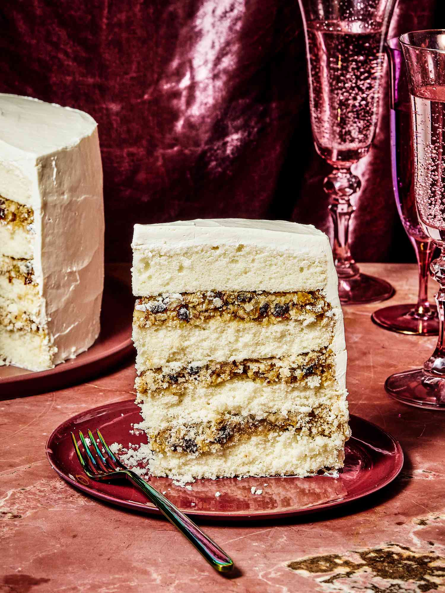 Alabama lane cake, with a slice placed on a red plate, on a red marble tabletop. 2 Magenta glasses of bubbly water, and a magenta fabric draped in the background. 