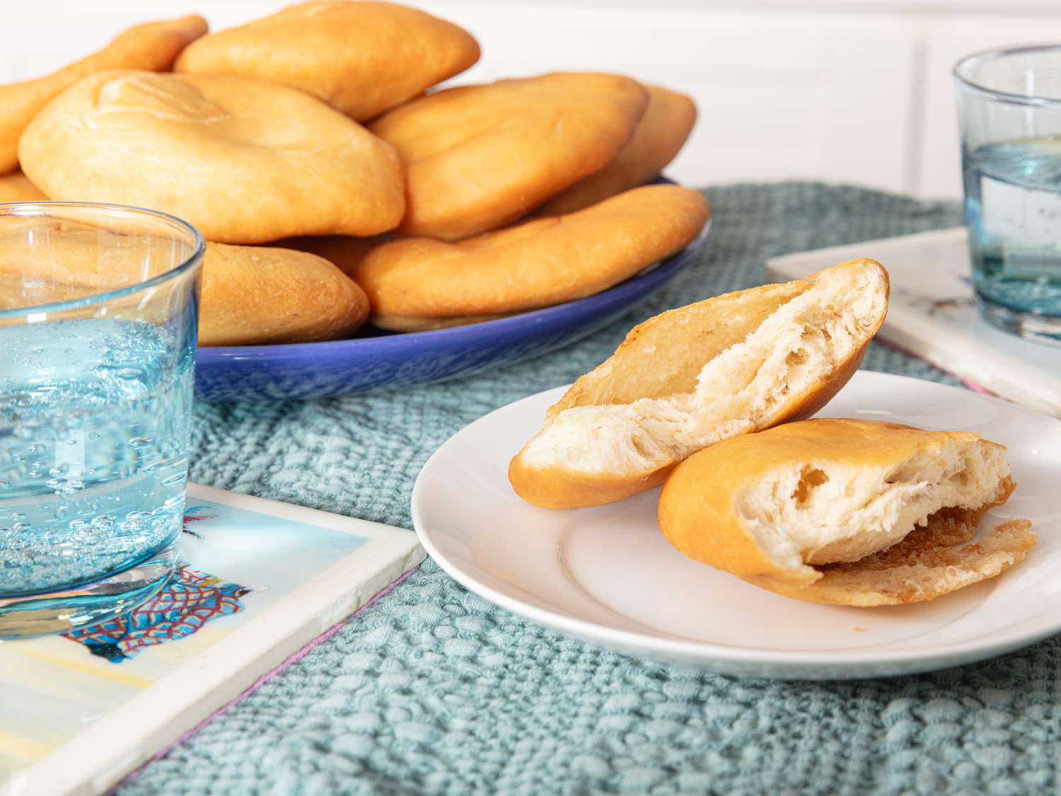 Platter of Johnny Cakes on a blue platter, with a cake on a smaller plate in front, ripped open. 2 glasses of sparkling water and blue tablecloth 