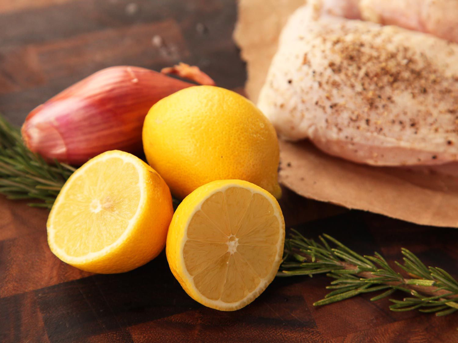 Close-up of ingredients for the recipe arranged on an an end-grain cutting board: a shallot, two lemons, a long sprig of rosemary, and seasoned chicken breasts.