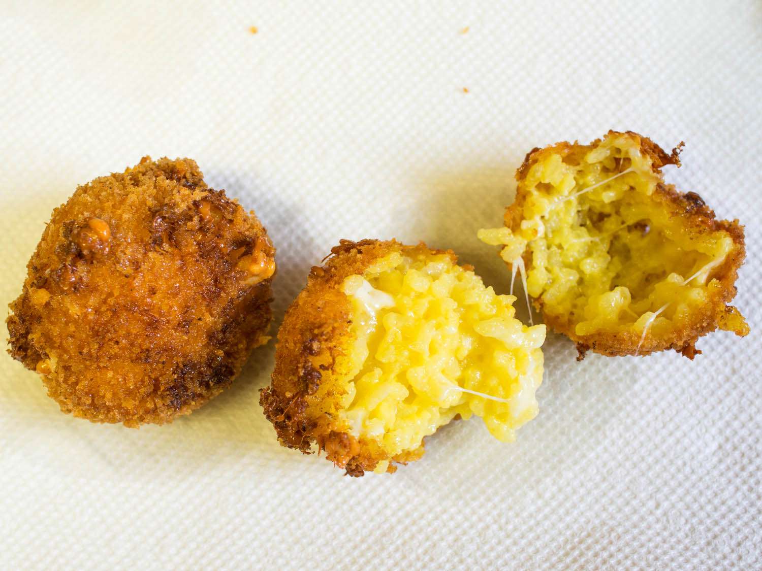 Close-up of two disheveled arancini draining on a paper towel. One is whole, unevenly browned with cheesy ruptures dotting its mottled surface. The other has been split apart to reveal shallow deposits of molten mozzarella near the outer crust.