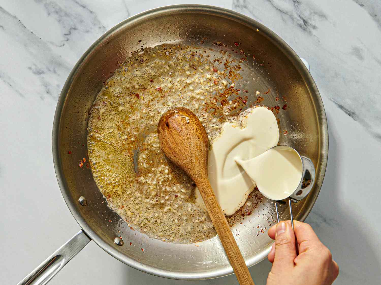 A hand pouring cream into a pan with sauted ingredients stirring with a wooden spoon