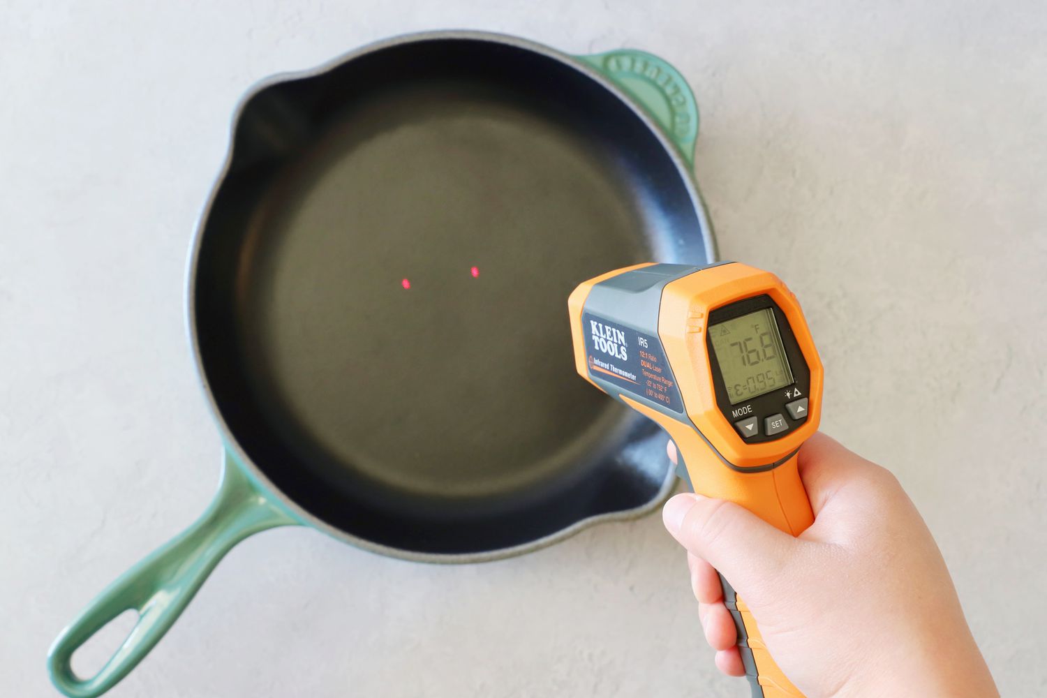 A person pointing an infrared thermometer at an enameled cast iron skillet with a dark-colored interior