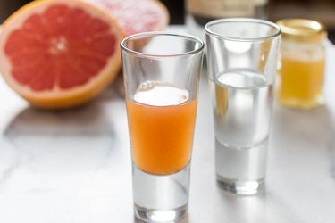 Closeup of a shot glass of grapefruit sangrita with chipotle and honey, flanked by another shot glass full of silver tequila. A halved grapefruit is in the blurred background.