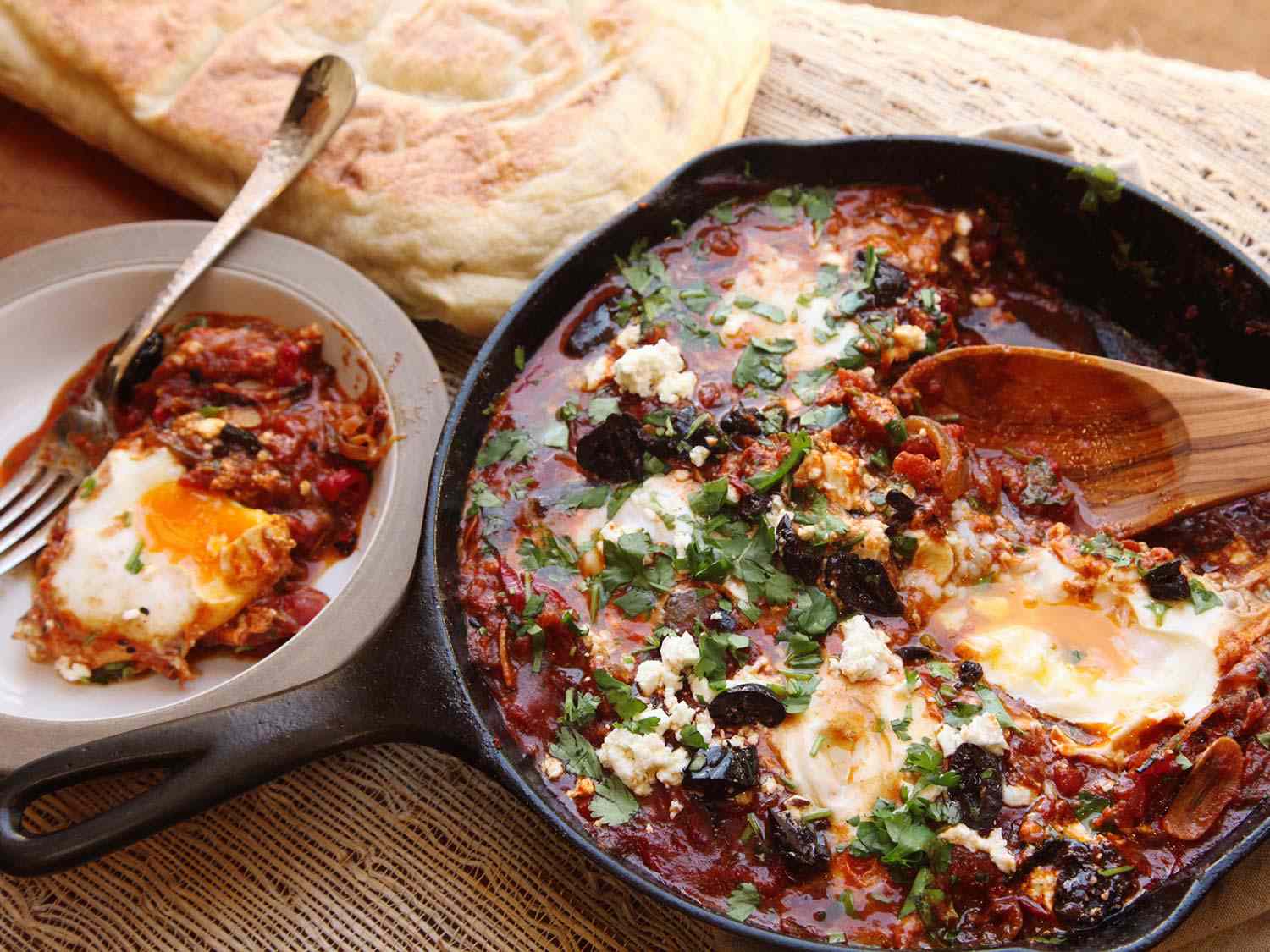 Shakshuka in a cast iron skillet with a serving in a small bowl on the side, with pita bread in the background.