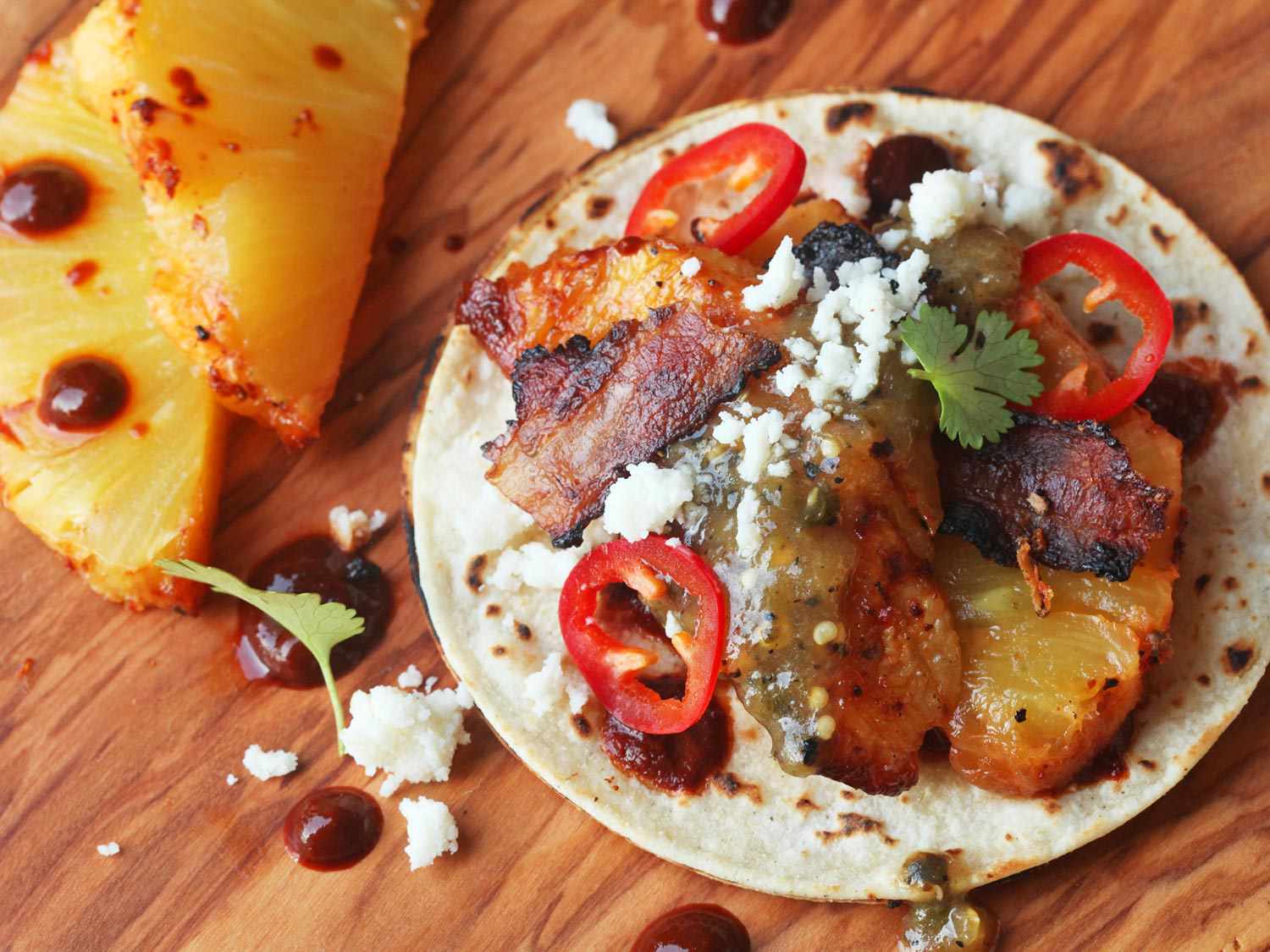 Closeup of a finished bacon-wrapped roasted pineapple taco, garnished with thin slices of red chile and crumbled cotija cheese.