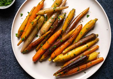 A round white porcelain plate holding multi-colored sous vide glazed carrots. The plate is on a dark blue textured surface and there is a small white bowl holding chopped parsley in the top left corner of the image.