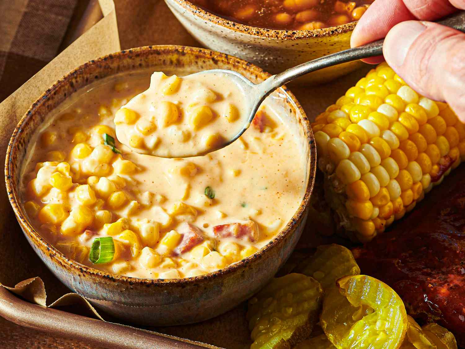 A bowl of cheesy corn garnished with diced vegetables served alongside a cob of corn pickle slices and barbecue items