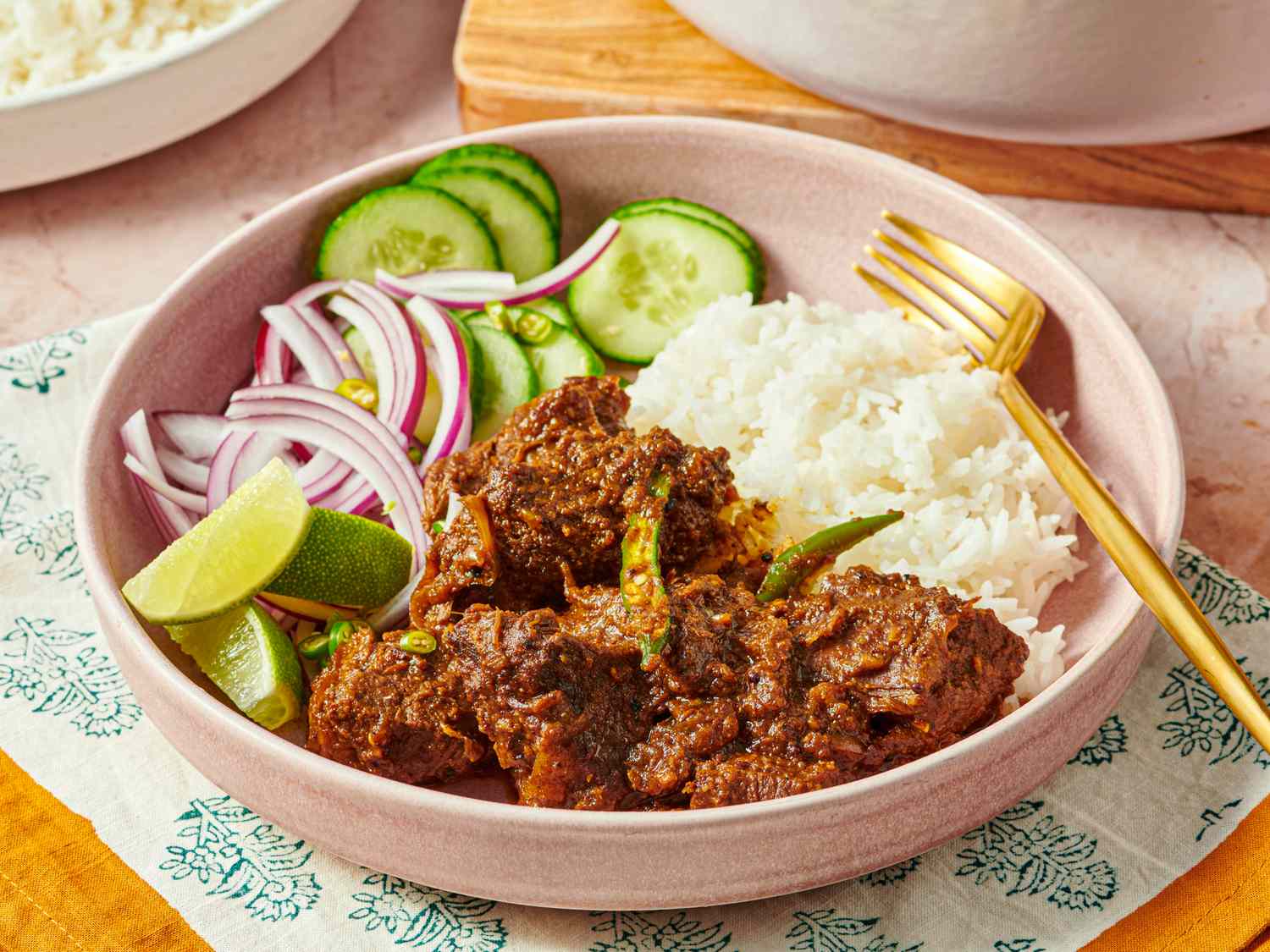 A bowl containing rice slices of cucumber and onion limes and a beef curry dish served with a fork