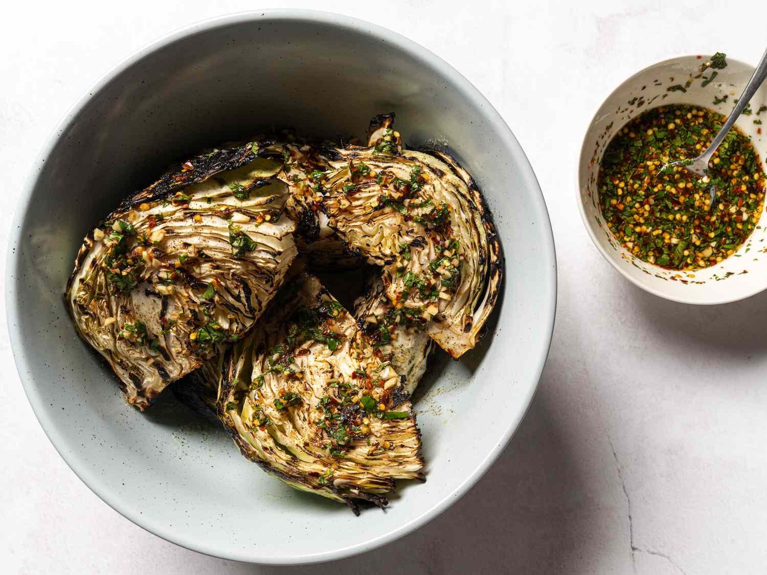 The grilled, sauce-drizzled cabbage wedges in a blue bowl. On the right hand side of the image is a small white bowl holding a spoon and additional sauce.