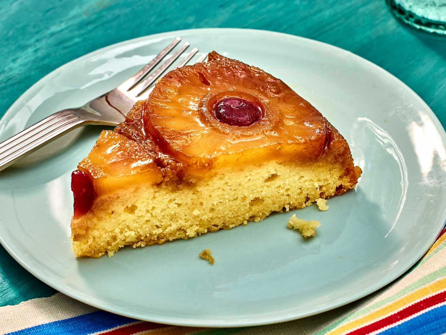 Side view of a slice of pineapple upside down cake with a fee loose crumbs, and a fork on a blue plate. The table cloth is a aqua color with stripes on it. 