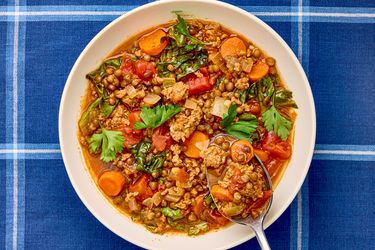 A bowl of sausage and lentil soup with vegetables garnished with parsley on a blue plaid tablecloth