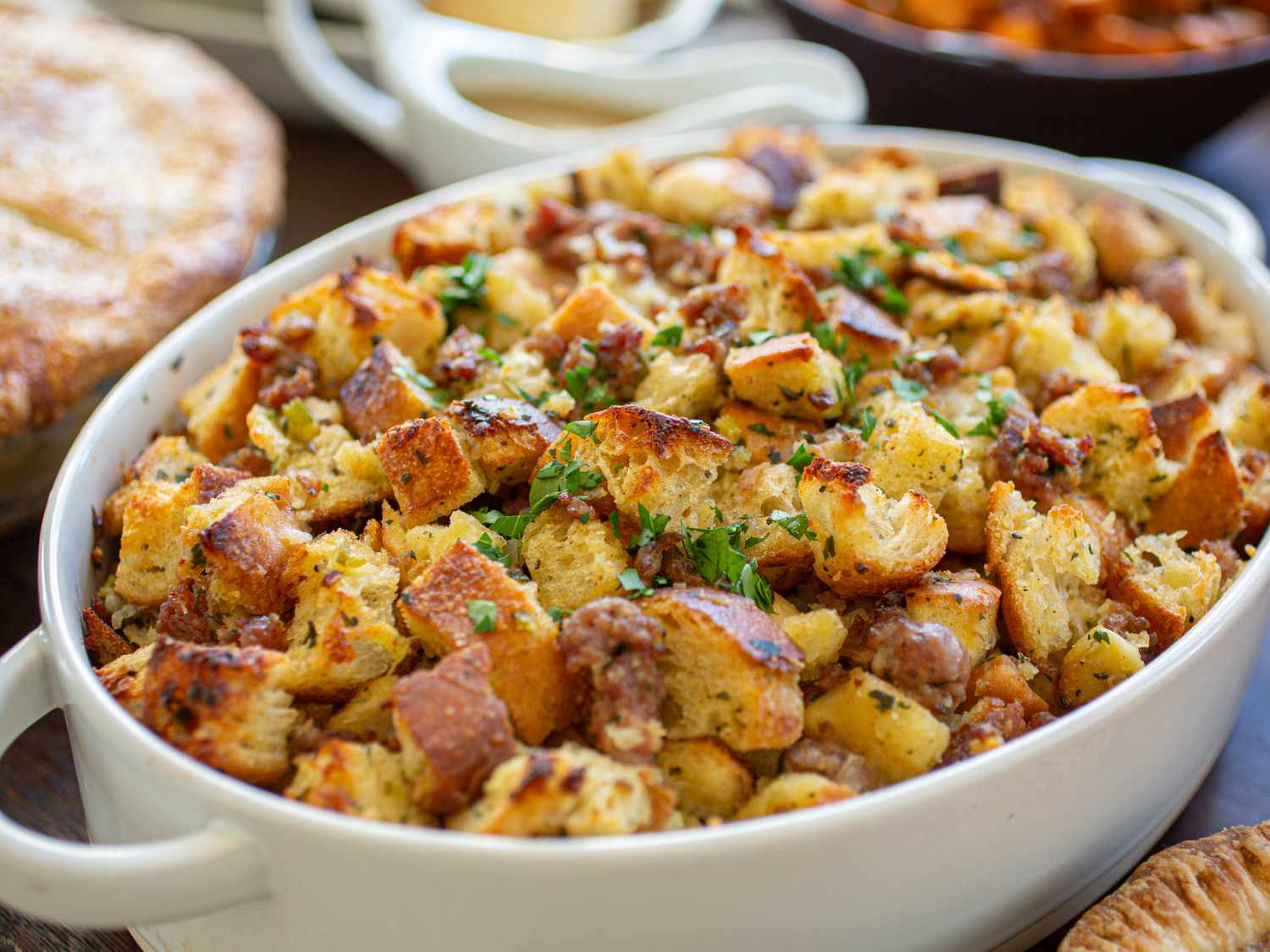 A dish of stuffing with herbs and bread cubes on a table with other dishes