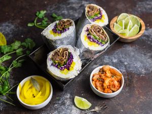 Korean beef bulgogi burritos, wrapped in foil, halved, and served as a "bouquet" in a loaf pan. Ramekins of kimchi, pickled daikon, and lime wedges are nearby.