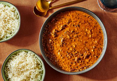 Dal Makhani in a bowl with 2 gold spoons, and 2 bowls of rice. Burnt orange colored surface and blue glass of water. 