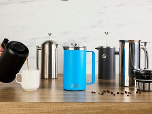 Five French press coffee makers on a wooden countertop. A person is pouring coffee from a French press into a mug.
