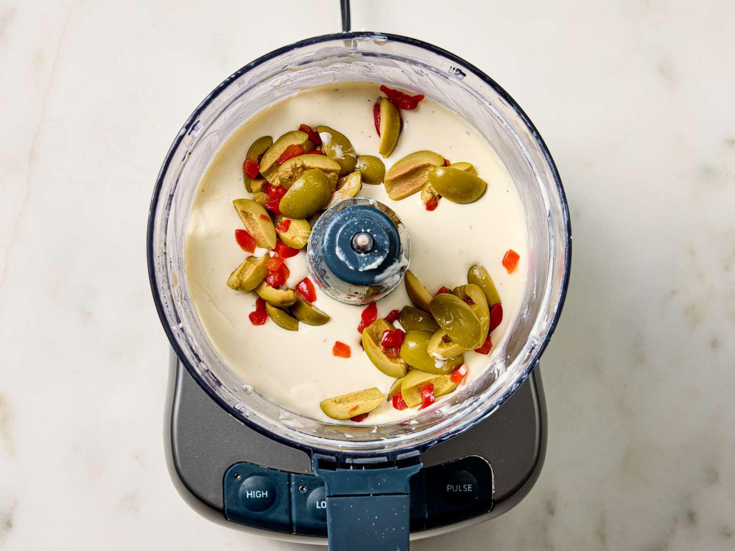 Food processor with creamy mixture and sliced peppers and olives on top