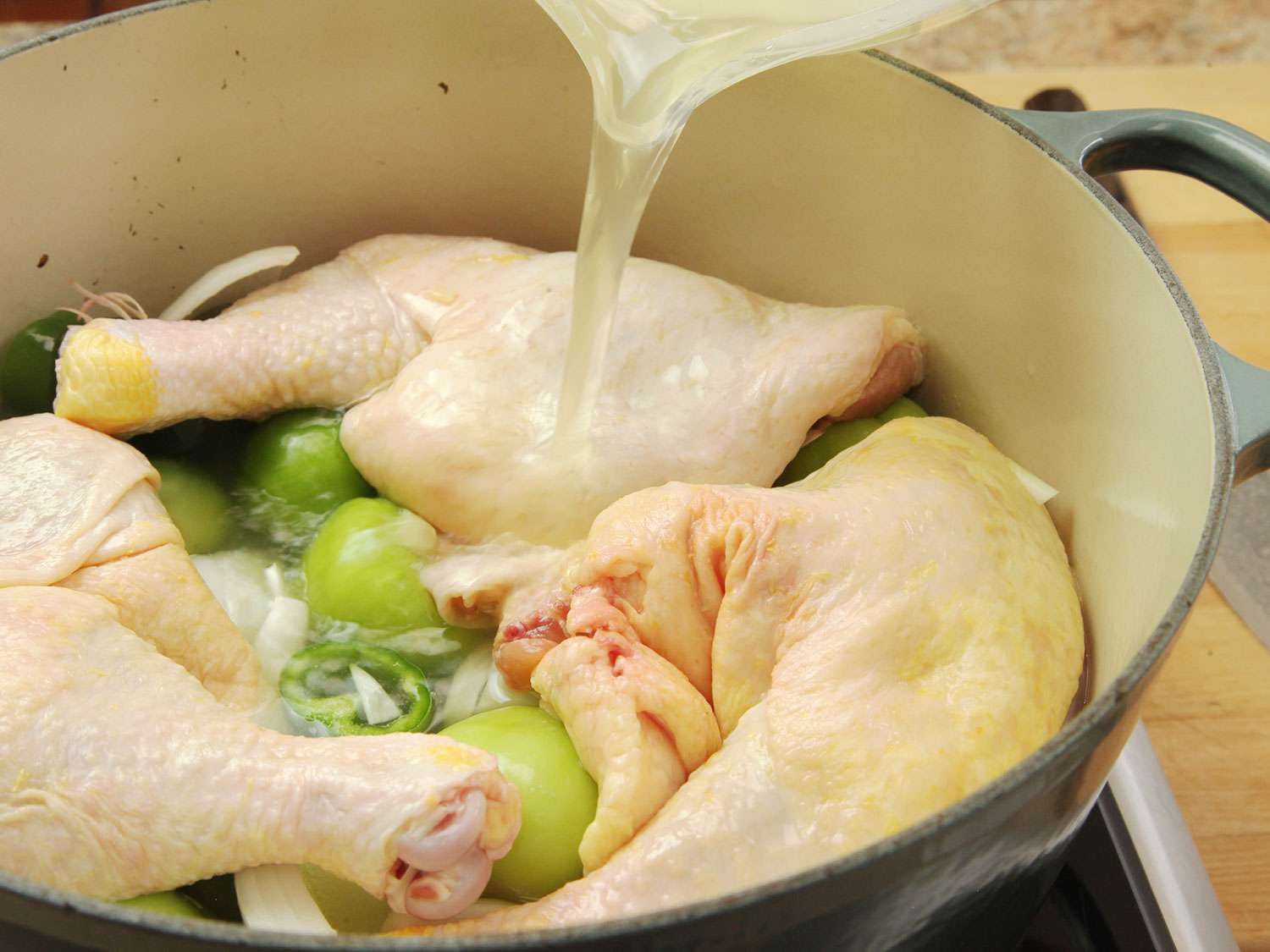 Chicken stock being poured over chicken leg quarters, which are layered on top of tomatillos, onions, and jalapeños in a Dutch oven for pozole.
