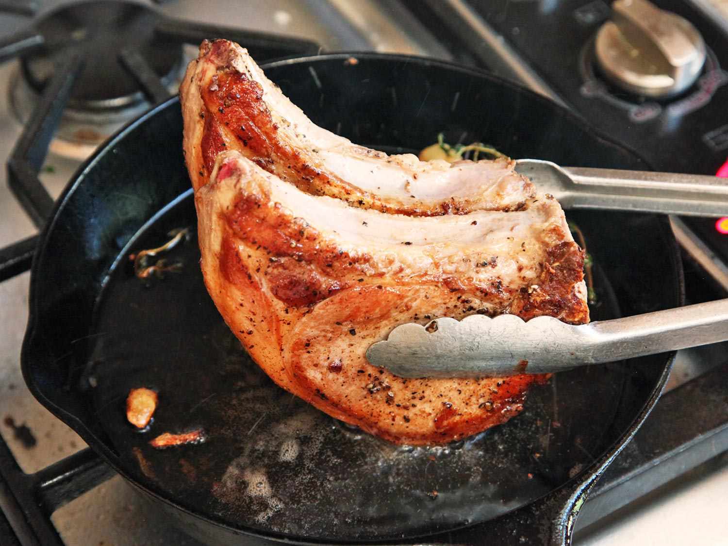 Two pork chops being picked up with tongs and having their sides cooked in a cast iron skillet.