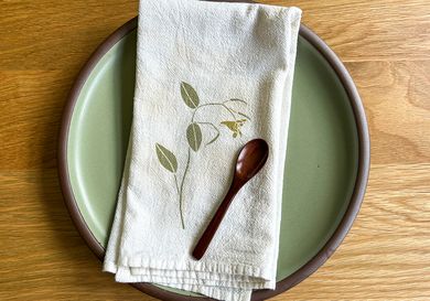 A ceramic plate with a cloth napkin and wooden spoon placed on top, featuring a leaf and flower pattern on the napkin