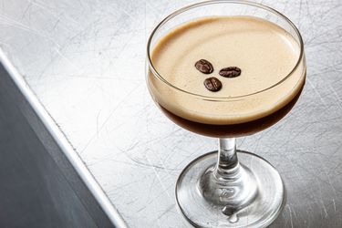 A glass of espresso martini garnished with three coffee beans on a metallic surface