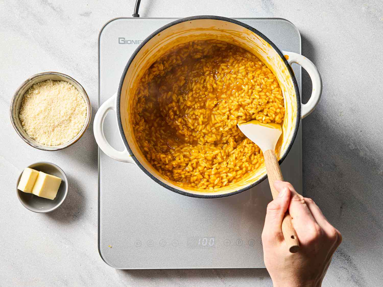 A person stirring a pot of carrot risotto on a stovetop, with parmesan and butter nearby