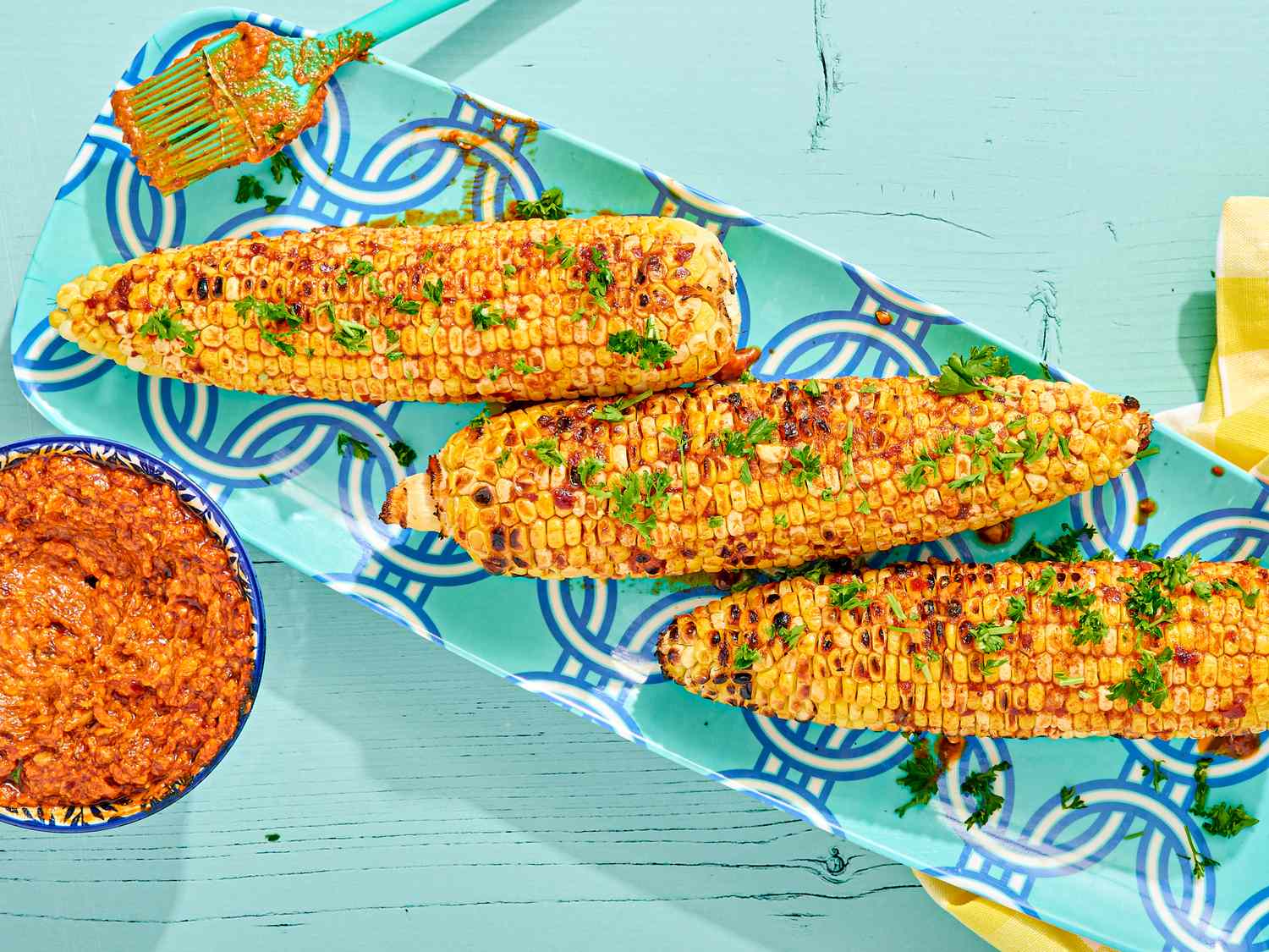 Grilled corn on a platter with parsley garnish, accompanied by a bowl of red sauce