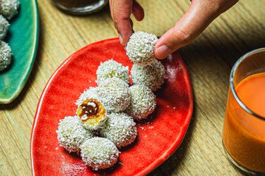 Overhead view of a hand picking up an onde-onde from a platter full with one cut open to see the inside 