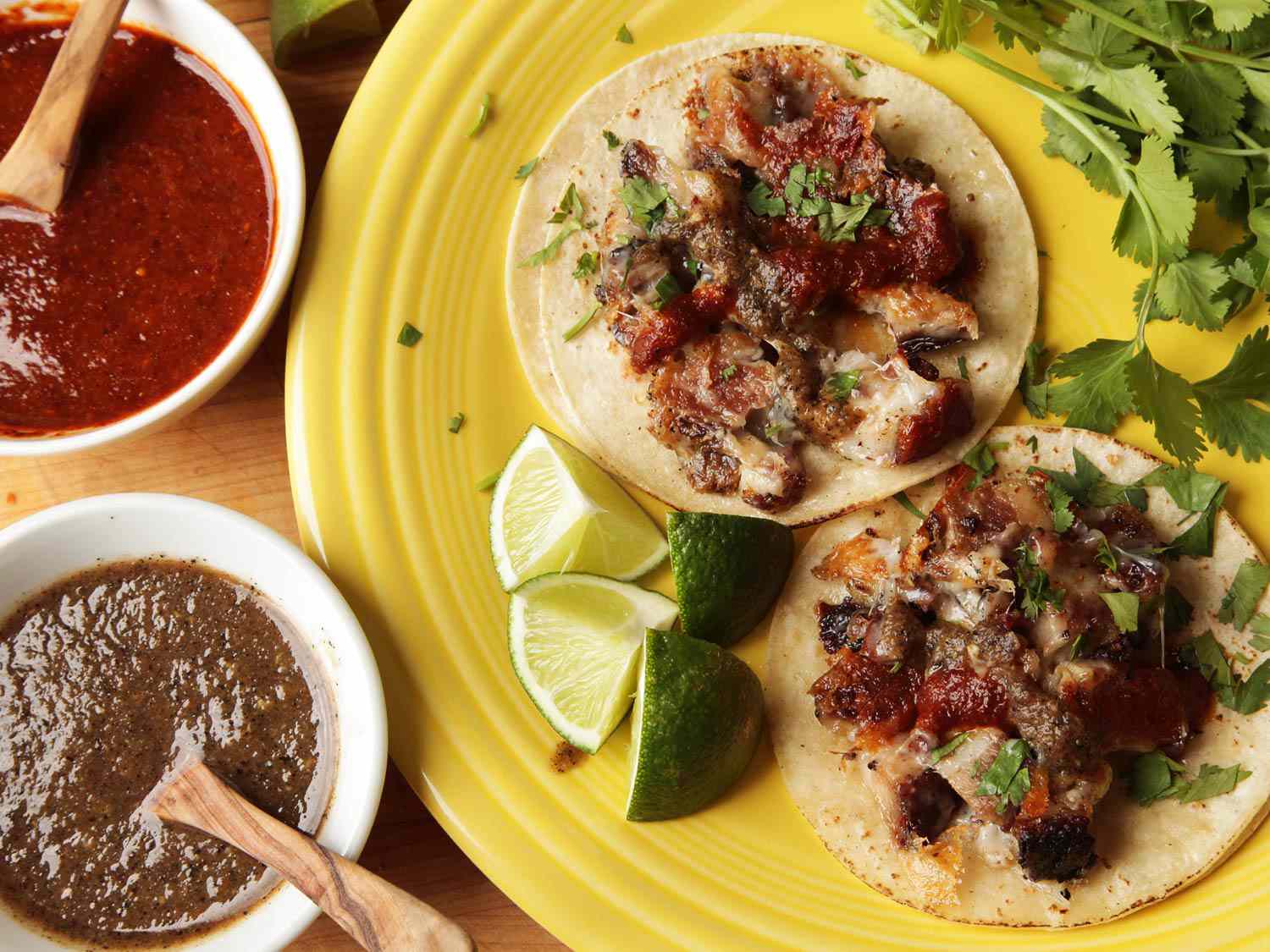 Overhead view of a plate of tacos de castacán, flanked by lime wedges, cilantro sprigs, and bowls of salsa.