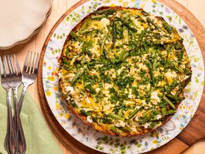 Smashed potato tart with asparagus and herbs on a floral plate, placed on a wooden table with forks and a green napkin nearby.