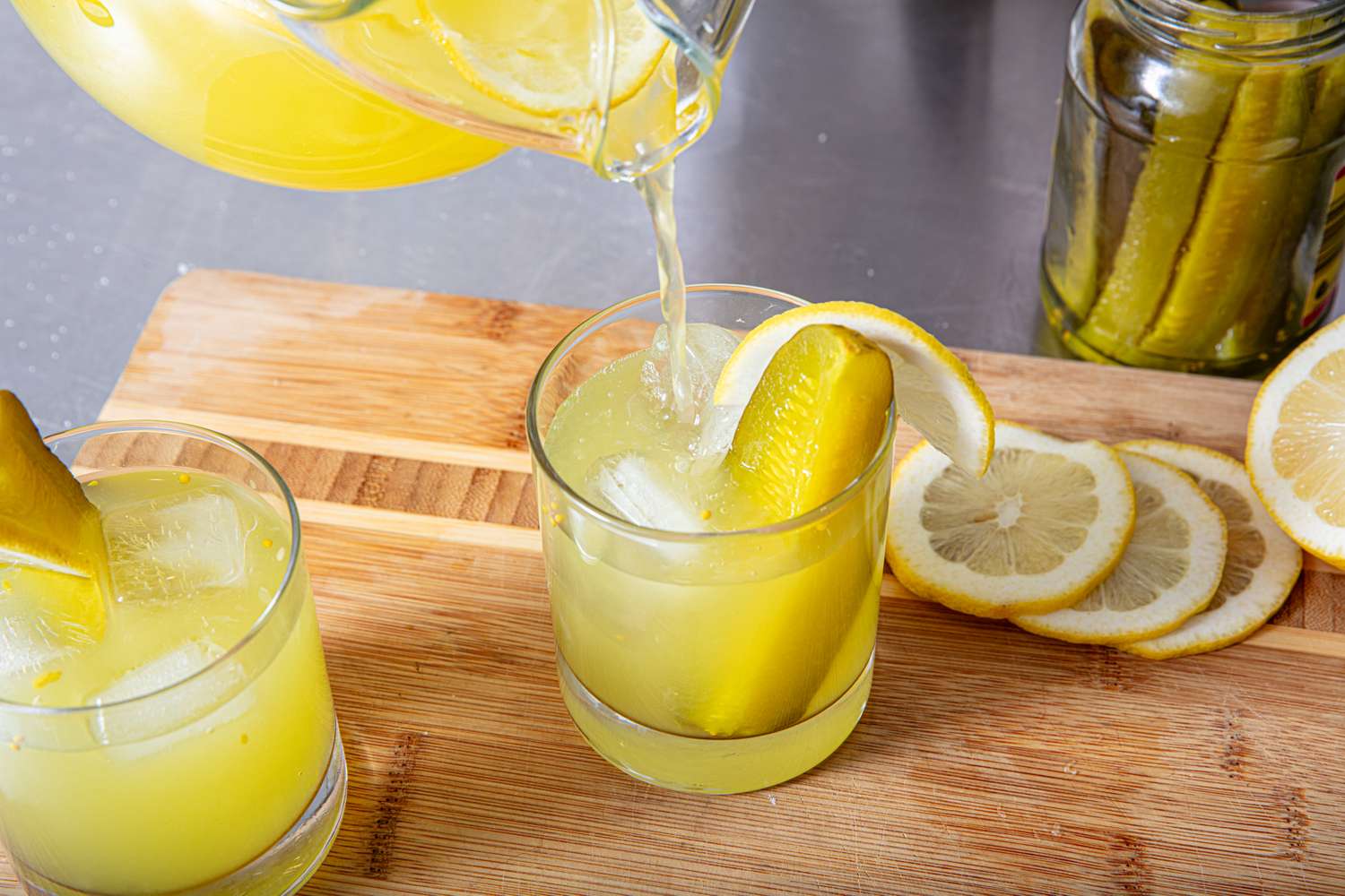 Glasses of lemonade garnished with pickle spears lemon slices and a pitcher pouring more lemonade