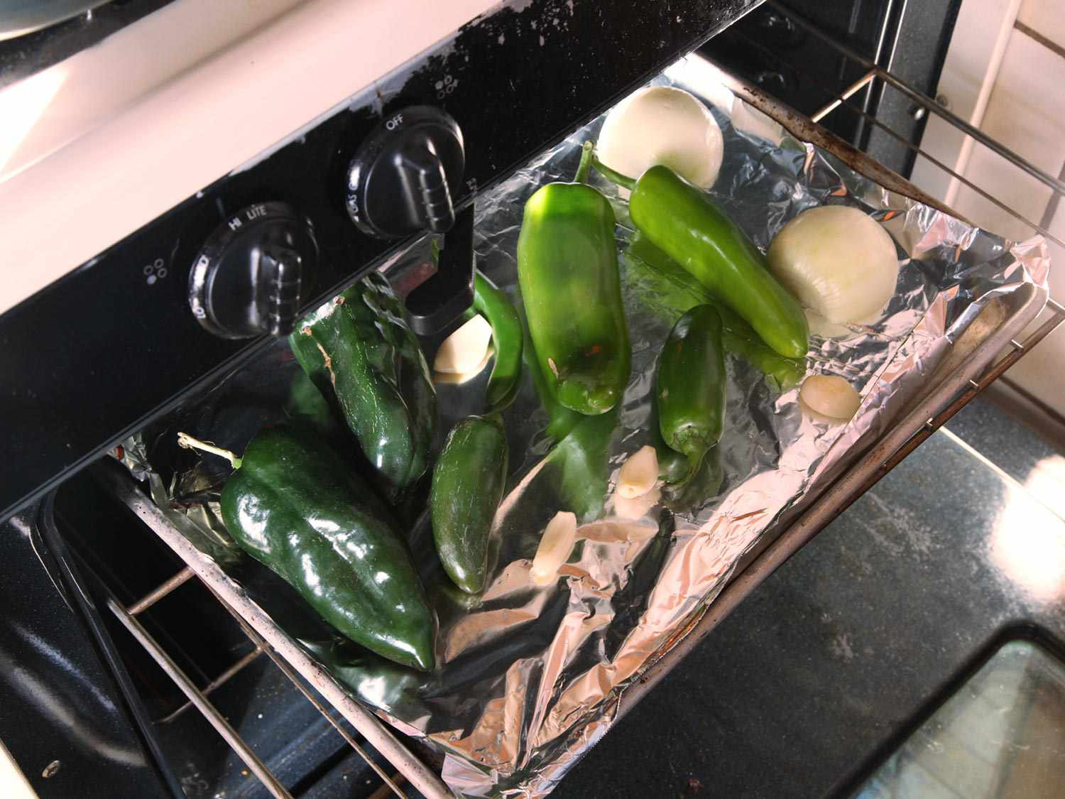 Foil-lined sheet pan of green chiles, onion, and garlic on an oven shelf.