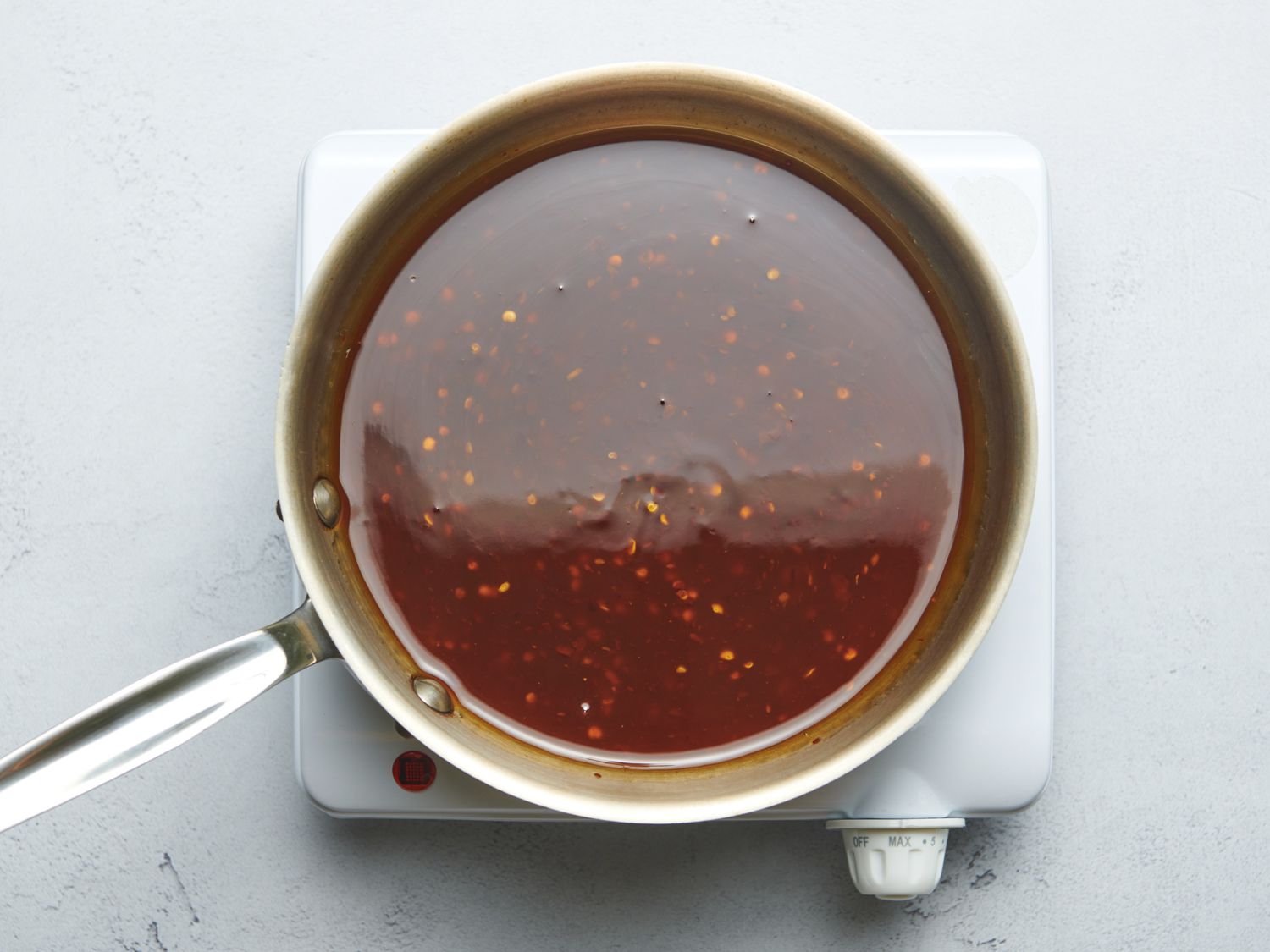 Sweet and sour sauce inside of a stainless steel saucepan after itâs stopped boiling, showing itâs glossy and fully cooked. The pan is on a small portable burner on a stone counter.
