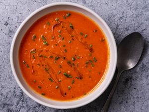 A bowl of creamy vegan tomato soup on a stone background with a spoon to the right of the bowl. The surface of the soup is drizzled with olive oil and sprinkled with chopped herbs.