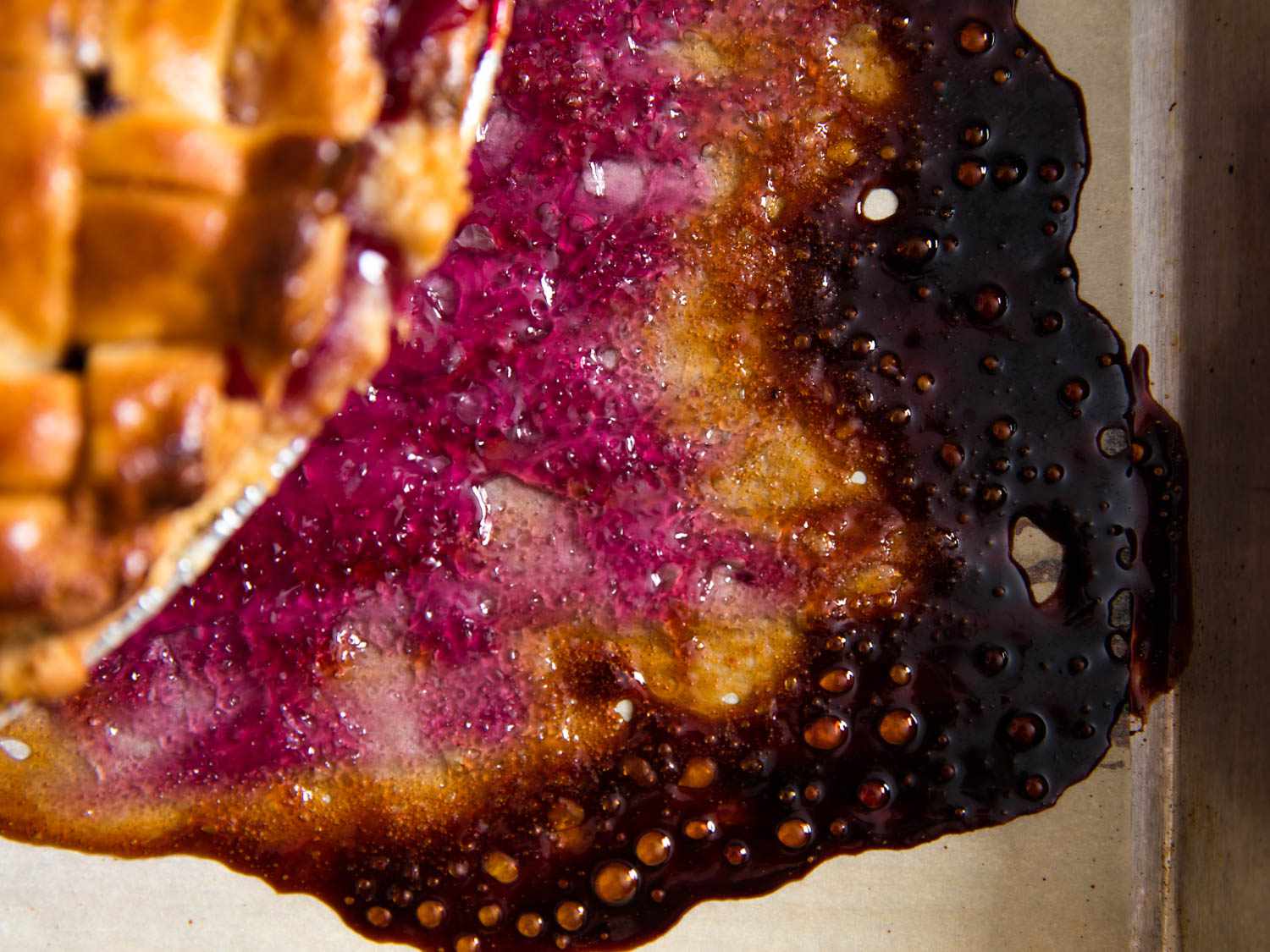 Overhead close-up of the baked puddle of fruit filling juice. The puddle has several distinct rings: an outer ring of completely browned filling juice; an intermediate ring of dried, caramelized filling juice; and, finally, a thick interior puddle of pinkish-purple filling juice.