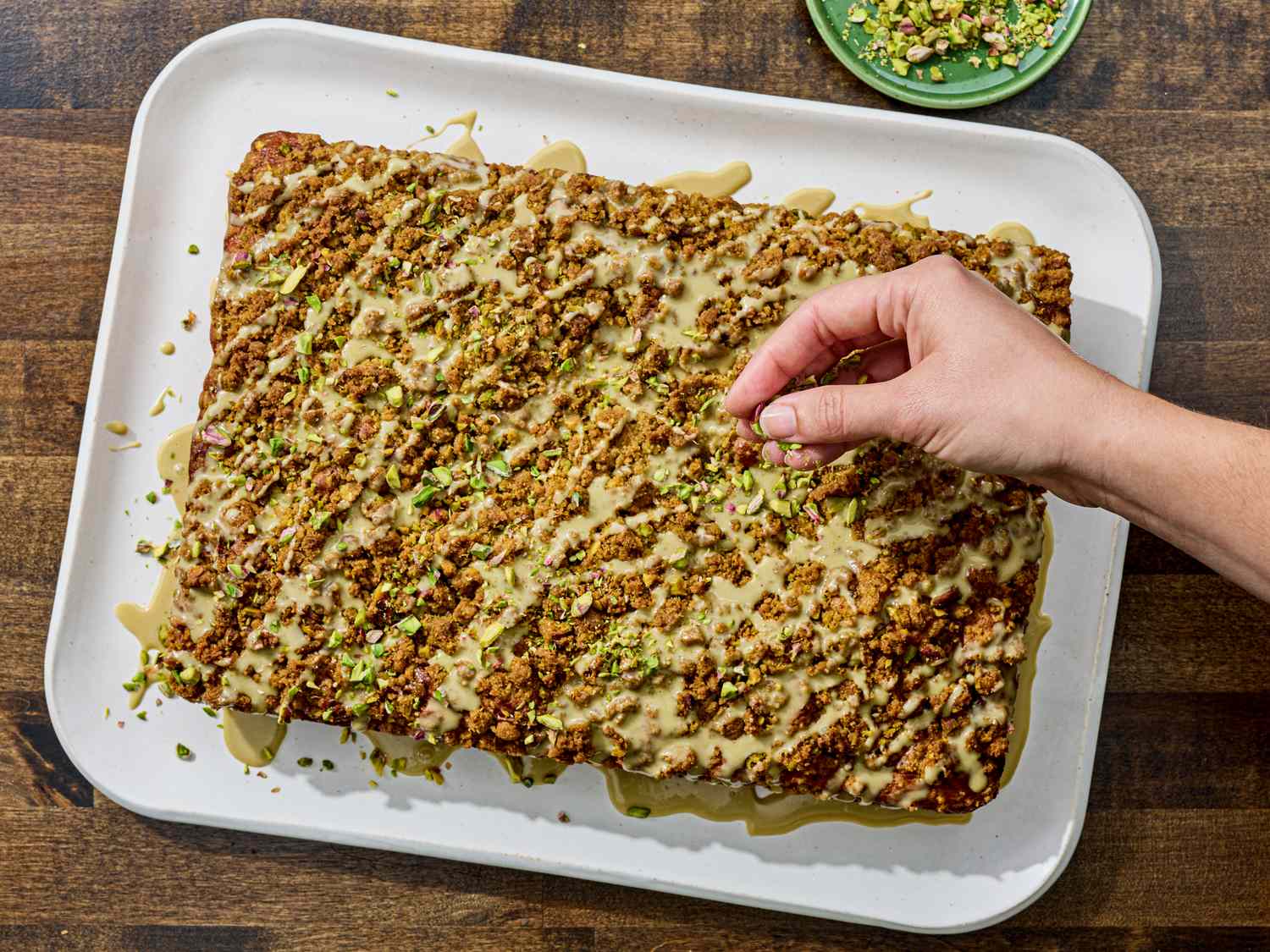 A hand sprinkling crushed pistachios onto a pistachio coffee cake on a tray