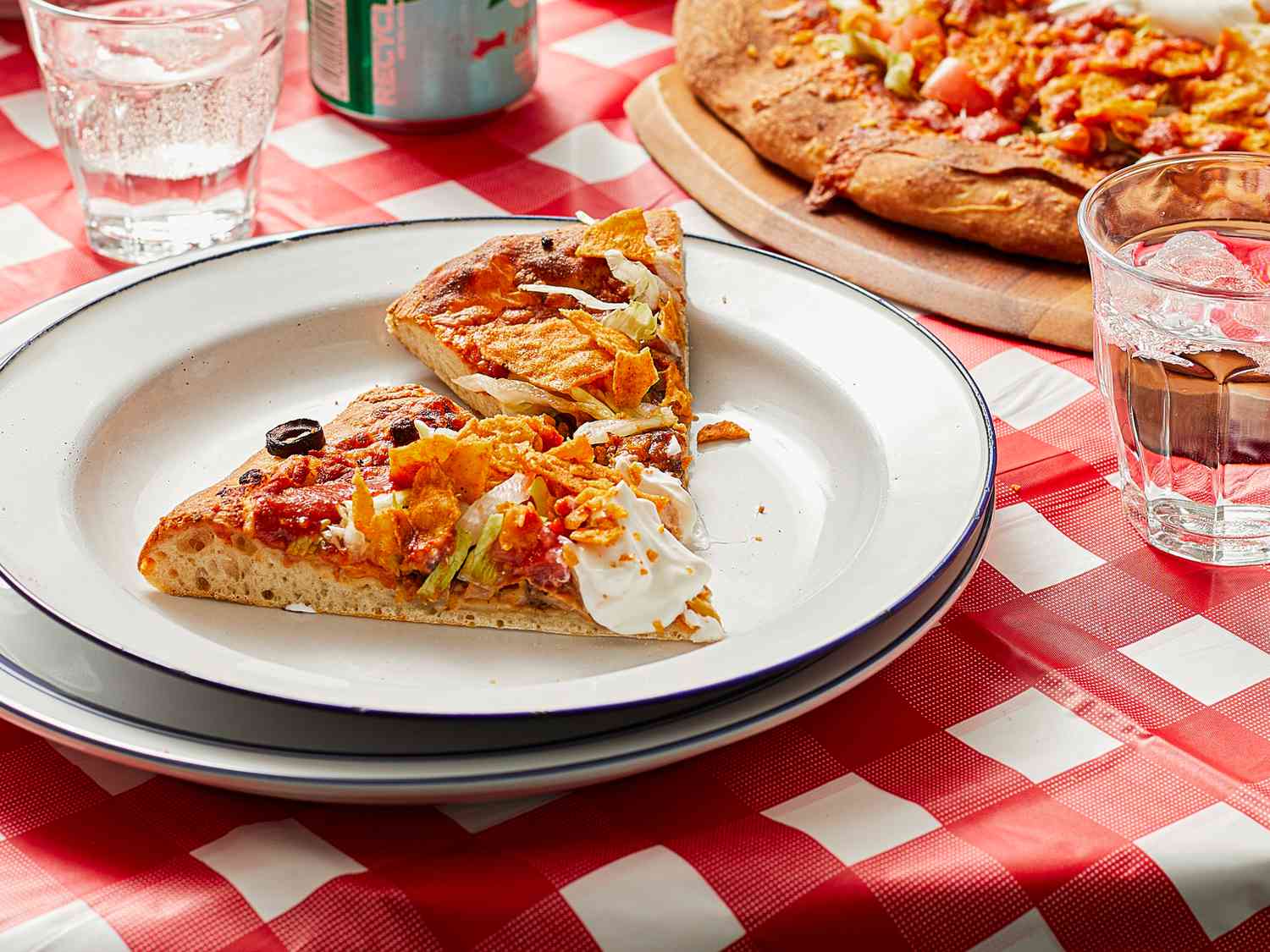 Two slices of taco pizza on a plate on a checkered tablecloth, a whole pizza and drinks in the background.