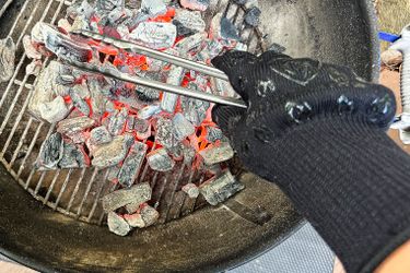 A hand in a heatresistant glove holding metal cooking tongs over smoldering charcoal in a grill